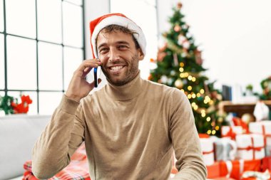 Genç İspanyol adam akıllı telefondan konuşuyor. Yılbaşı ağacının yanında oturuyor.