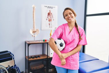 Young hispanic woman working at physiotherapy clinic holding football ball looking positive and happy standing and smiling with a confident smile showing teeth 