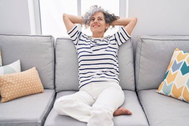 Orta yaşlı, kır saçlı, elleri başının üstünde, evinde kanepede oturan bir kadın.