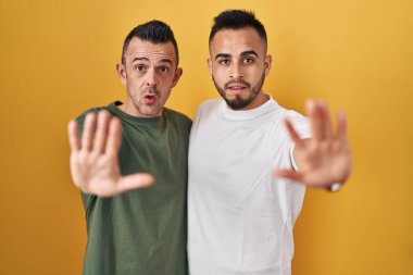 Homosexual couple standing over yellow background doing stop gesture with hands palms, angry and frustration expression 