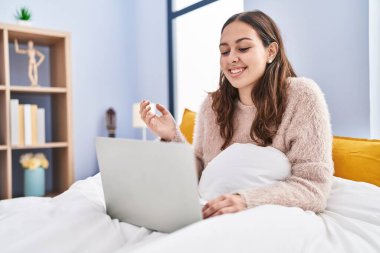 Young hispanic woman using computer laptop on the bed screaming proud, celebrating victory and success very excited with raised arm 