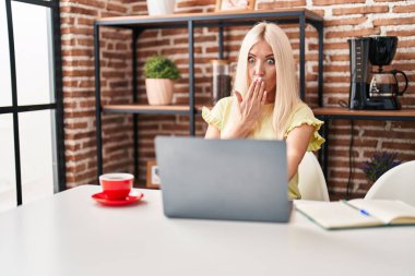 Caucasian woman doing video call with laptop covering mouth with hand, shocked and afraid for mistake. surprised expression 
