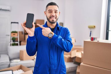Hispanic man working on moving service showing smartphone screen smiling happy pointing with hand and finger 