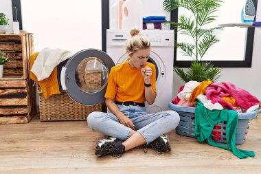 Young blonde woman doing laundry sitting by washing machine feeling unwell and coughing as symptom for cold or bronchitis. health care concept. 