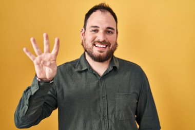 Plus size hispanic man with beard standing over yellow background showing and pointing up with fingers number four while smiling confident and happy. 