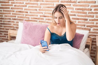 Young blonde woman using smartphone with surprise expression at bedroom