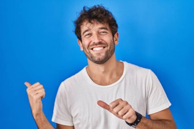 Hispanic young man standing over blue background pointing to the back behind with hand and thumbs up, smiling confident 