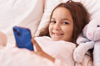 Adorable hispanic girl using smartphone lying on bed at bedroom