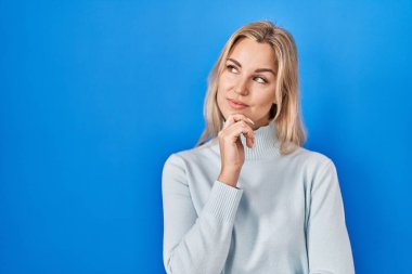 Young caucasian woman standing over blue background with hand on chin thinking about question, pensive expression. smiling with thoughtful face. doubt concept. 