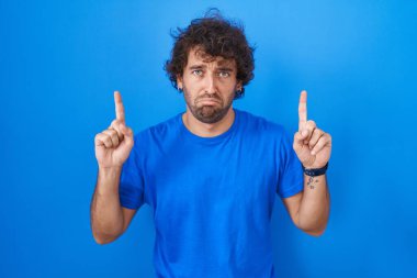 Mavi arka planda dikilen İspanyol genç adam üzgün ve üzgün görünüyor, parmaklarıyla yön gösteriyor, mutsuz ve depresif.. 