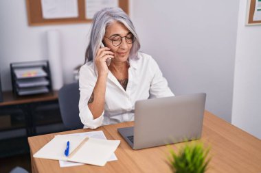 Orta yaşlı, kır saçlı, iş kadını. Ofiste bilgisayarla konuşuyor.