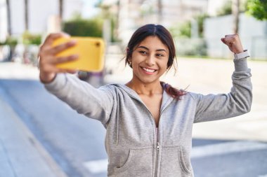 Young hispanic woman make selfie doing strong gesture at street