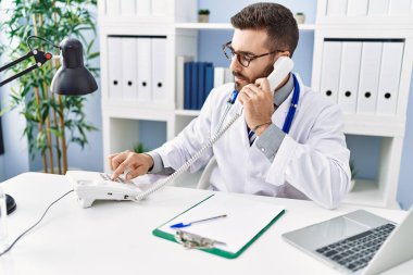 Doktor üniforması giyen İspanyol genç adam klinikte telefonla konuşuyor.