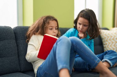 Two kids reading book sitting on sofa at home