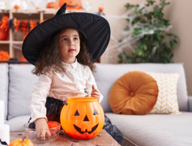 Adorable hispanic girl having halloween party putting sweets on pumpkin basket at home