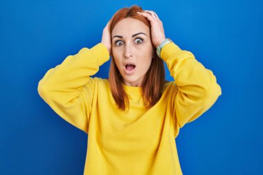 Young woman standing over blue background crazy and scared with hands on head, afraid and surprised of shock with open mouth 