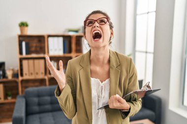 Young woman working at consultation office crazy and mad shouting and yelling with aggressive expression and arms raised. frustration concept. 
