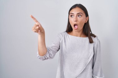 Young hispanic woman standing over white background pointing with finger surprised ahead, open mouth amazed expression, something on the front 