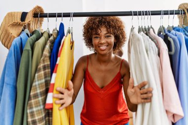 Young african american woman smiling confident appearing through rack at clothing store