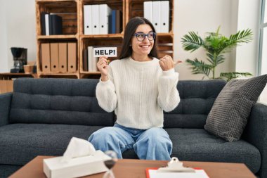 Young hispanic woman going to therapy at consultation office asking for help pointing thumb up to the side smiling happy with open mouth 
