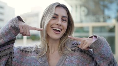 Young blonde woman smiling confident pointing with fingers to the mouth at street