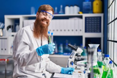 Young redhead man scientist holding test tubes writing report working at laboratory