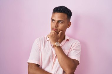 Pembe arka planda, elleri çenesinde, soru sormayı düşünen genç bir İspanyol. Düşünceli bir ifade. Gülümseyen ve düşünceli bir yüz. şüphe kavramı. 