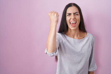 Young brunette woman standing over pink background angry and mad raising fist frustrated and furious while shouting with anger. rage and aggressive concept. 