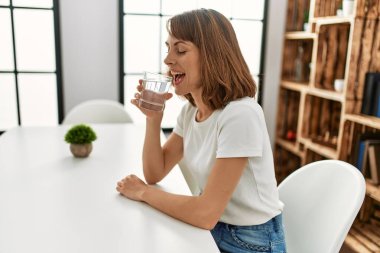 Beyaz kadın su içiyor. Evdeki masada oturuyor.