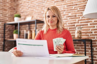 Genç sarışın kadın elinde dolarlarla evde doküman okuyor.