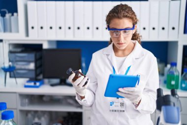 Young beautiful hispanic woman scientist using touchpad holding bottle at laboratory
