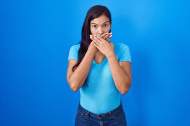 Mavi arka planda dikilen genç İspanyol kadın yanlışlıkla elleri ile ağzını kapatırken şok oldu. gizli kavram. 