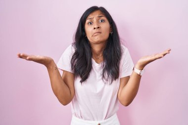 Pembe arka planda duran, elleri ve kolları havada, kafası karışmış genç İspanyol kadın. şüphe kavramı. 