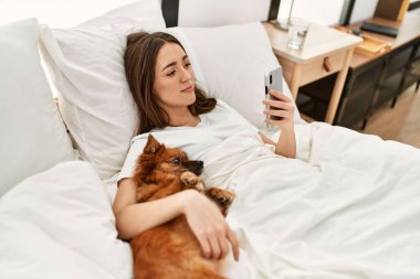 Genç İspanyol kadın köpeğe sarılıyor ve yatakta akıllı telefon kullanıyor.