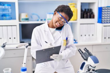 Bilim adamı üniforması giyen Afrikalı Amerikalı kadın. Akıllı telefondan konuşuyor. Laboratuvarda panoya yazı yazıyor.