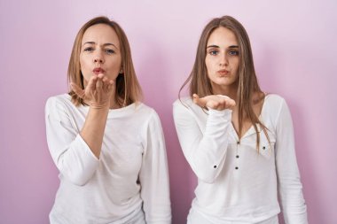 Middle age mother and young daughter standing over pink background looking at the camera blowing a kiss with hand on air being lovely and sexy. love expression. 