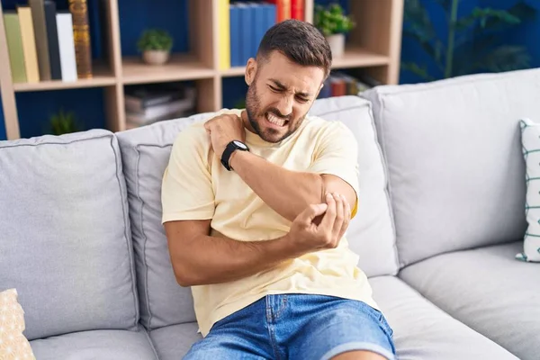 Young hispanic man sitting on sofa suffering for elbow pain at home