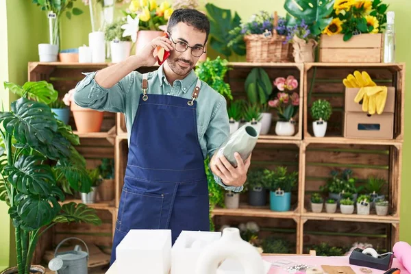 Young hispanic man florist talking on smartphone holding plant pot at flower shop