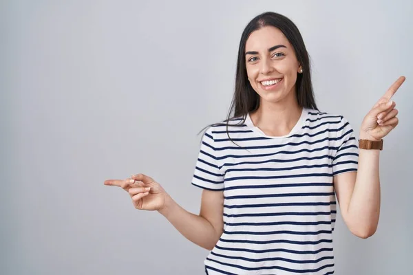 若いブルネットの女性は 異なる方向に指で自信を持って指して笑顔ストライプTシャツを着ています 広告のコピースペース — ストック写真
