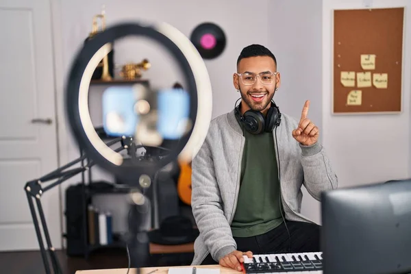 Young hispanic man playing piano at music studio recording himself smiling with an idea or question pointing finger with happy face, number one