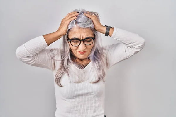 Middle age woman with grey hair standing over white background suffering from headache desperate and stressed because pain and migraine. hands on head.