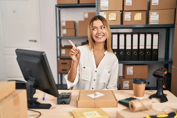 Young blonde woman working at small business ecommerce cheerful with a smile of face pointing with hand and finger up to the side with happy and natural expression on face