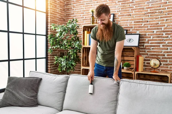 Young redhead man cleaning sofa using pet hair roller at home