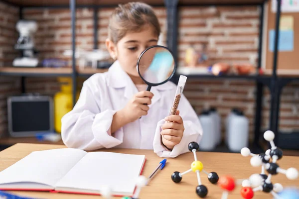 Adorable hispanic girl student looking test tube using magnifying glass at laboratory classroom