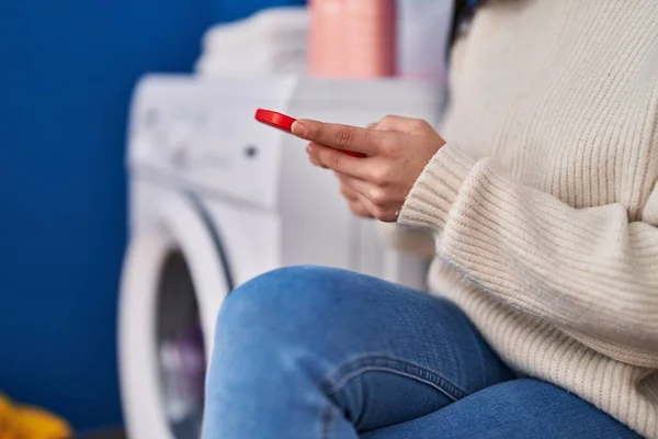 洗濯室で洗濯機を待っているスマートフォンを使用して若い美しいヒスパニック系の女性 — ストック写真