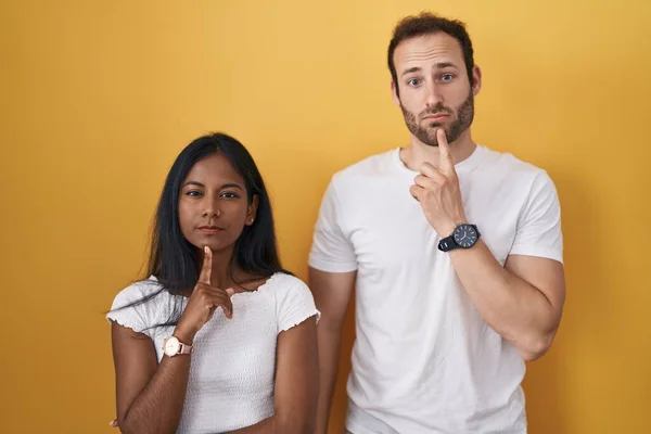 Interracial couple standing over yellow background thinking concentrated about doubt with finger on chin and looking up wondering