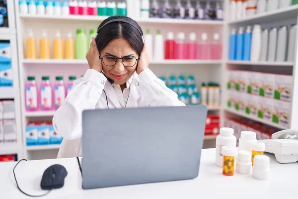 Young arab woman working at pharmacy drugstore using laptop covering ears with fingers with annoyed expression for the noise of loud music. deaf concept.