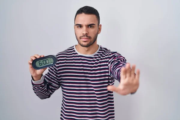 Ung Latinamerikan Man Bär Pyjamas Håller Väckarklocka Med Öppen Hand — Stockfoto
