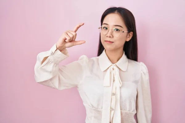 Young chinese woman standing over pink background smiling and confident gesturing with hand doing small size sign with fingers looking and the camera. measure concept.