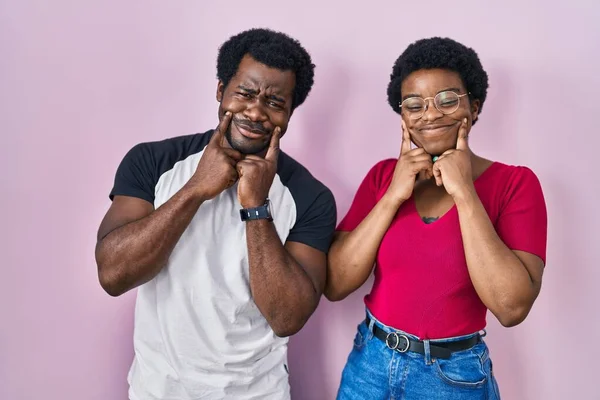 年轻的非洲裔美国夫妇站在粉红的背景上微笑着 张开嘴 指指点点着 强迫着欢乐的微笑 — 图库照片